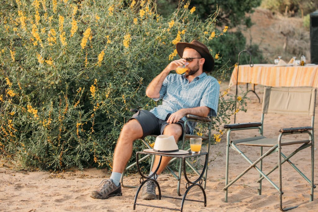 A man sits in a folding chair on the sand with a drink.