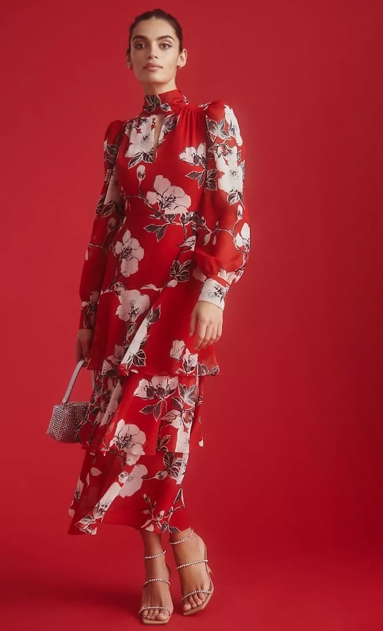 A young white model wears a long mock-neck red dress with a white floral pattern. She's posing against a red background.