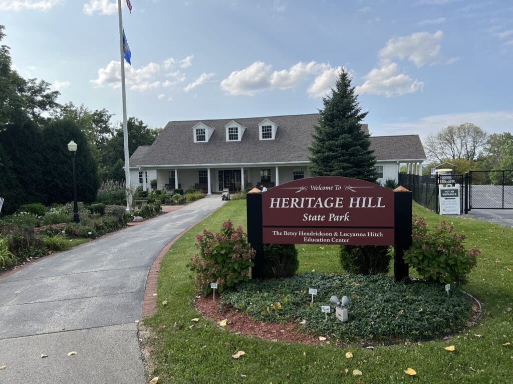 A two-story white house serves as the welcome center at Heritage Hill State Park in Green Bay, Wisconsin. The house features a long driveway with green grass and a brown Heritage Hill sign at the entrance. 