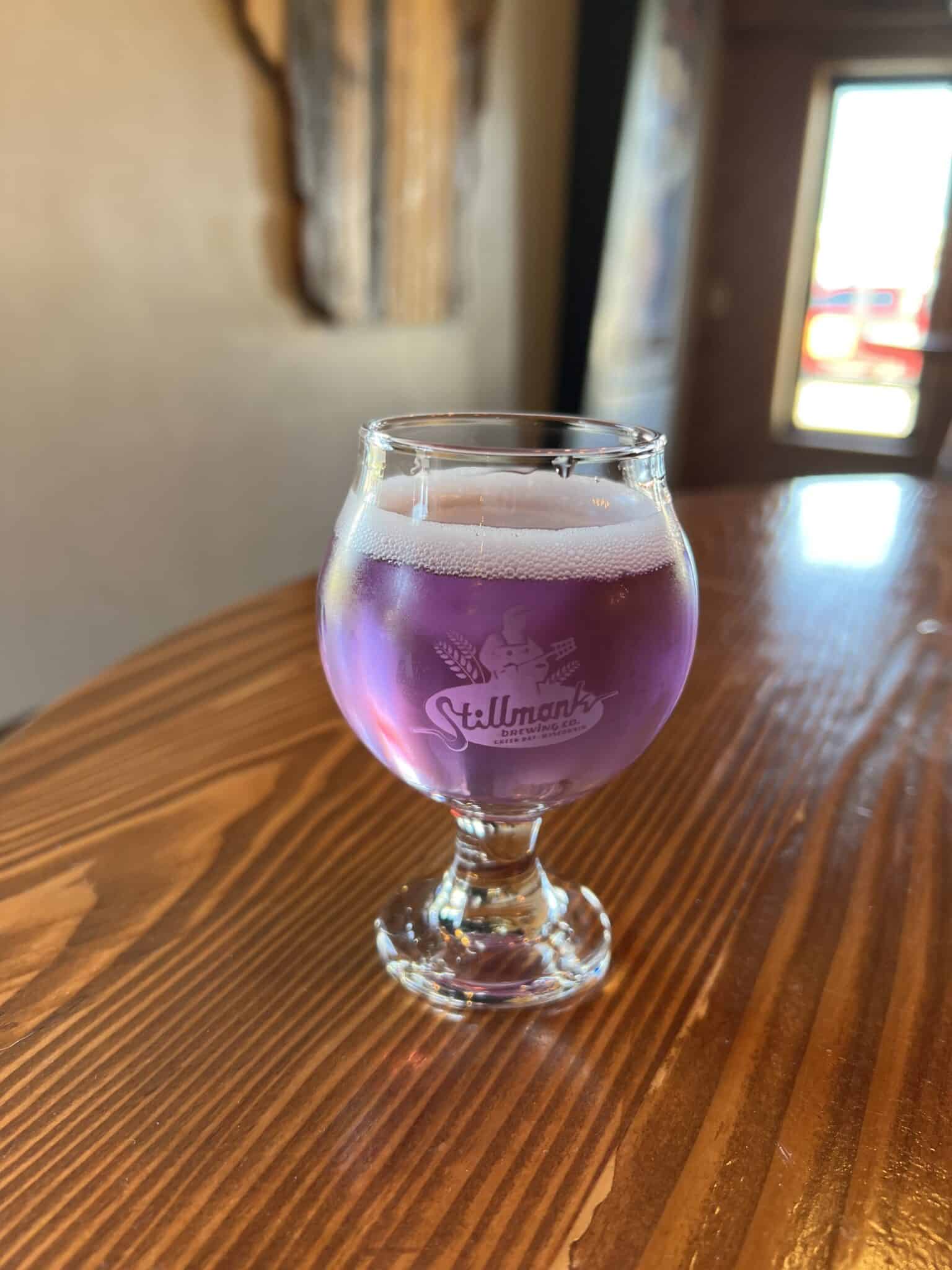 A clear goblet etched with the words Stillmark sits on a wooden bar and contains a purple-colored liquid. 