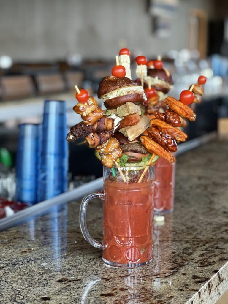 Two mugs atop a brown marble countertop are filled with red liquid. The Bloody Mary cocktails are spectacularly decorated with skewers of meat, cheese, pickled vegetables, and cherry tomatoes.