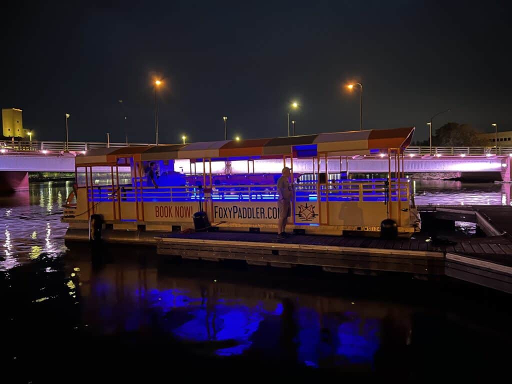 A flat, white peddle pontoon boat reads "Book Now! FoxyPaddler.com" on the side. The boat is shown in the water at night, lit by a blue light. 