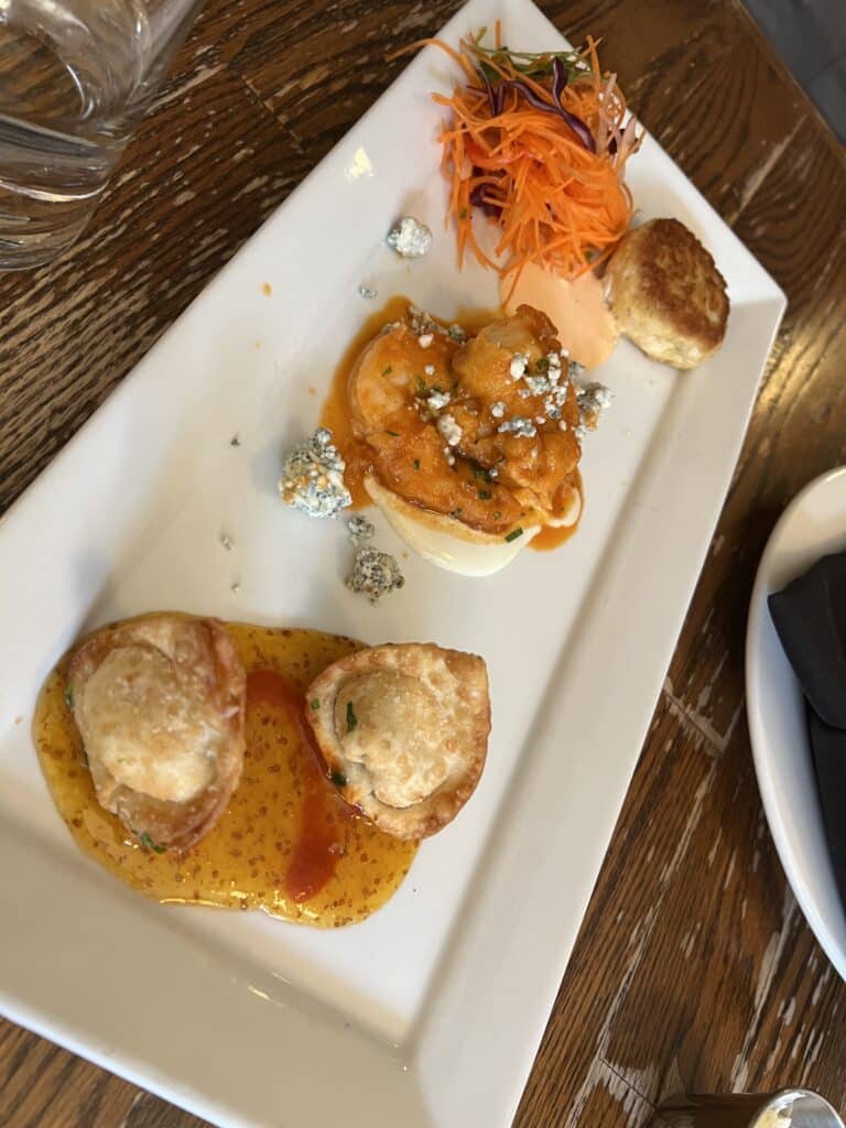 A trio of crabcake appetizers is served on a rectangular plate atop a wooden plank table. The appetizers are garnished and professionally plated. 