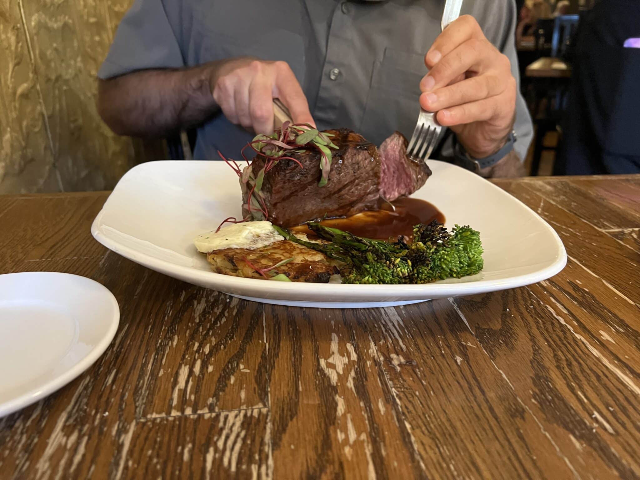 A person is shown cutting a steak on a white square plate atop a wooden table. The steak is professionally plated with vegetables and sauce.