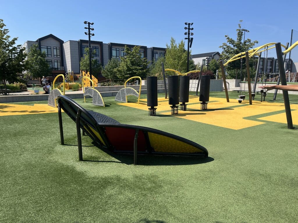A green grass-covered playground features several different climbing activities atop yellow mats. The climbing and playground equipment is black and silver with yellow accents. 