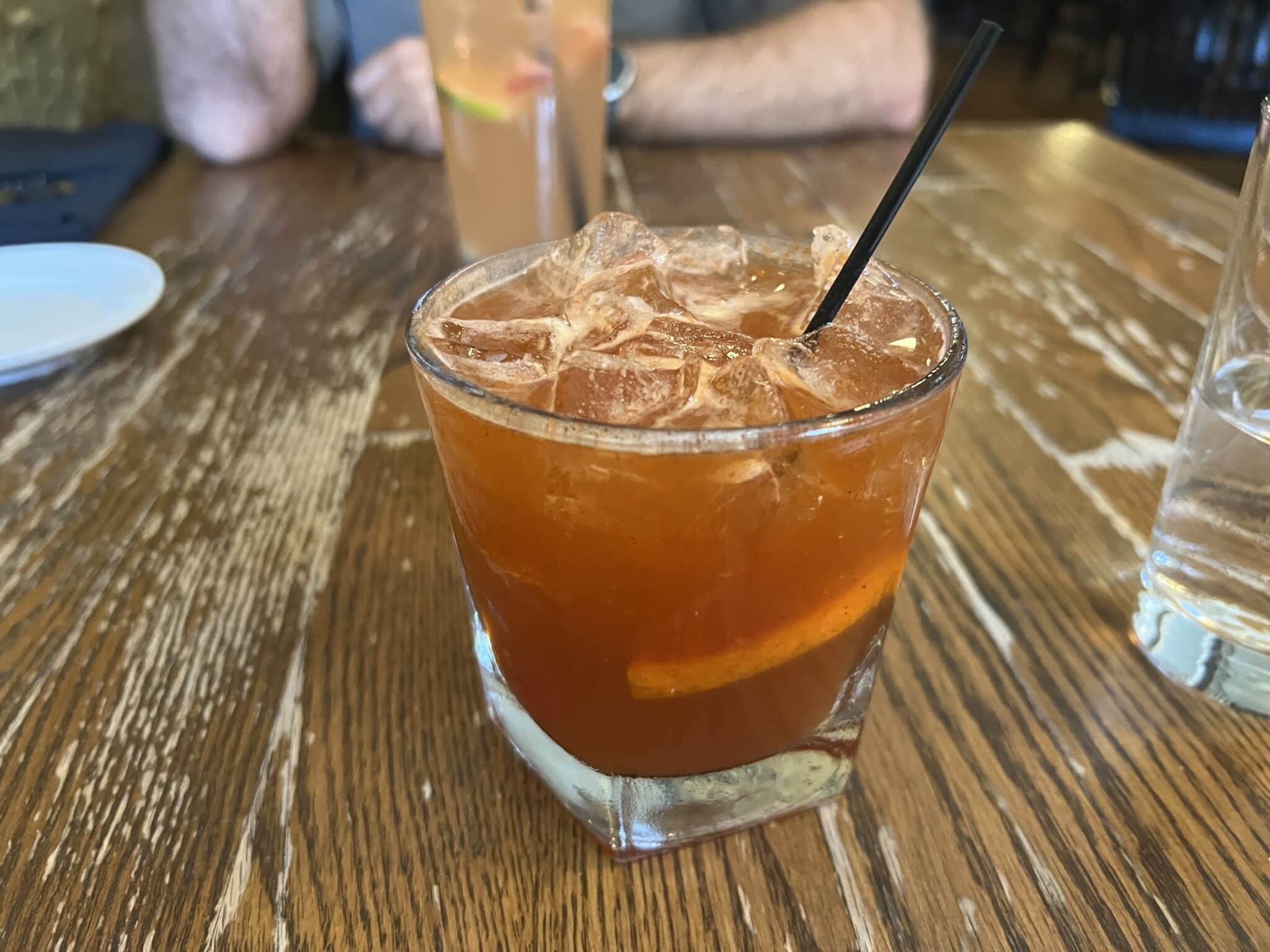 A brandy old-fashioned cocktail is served with ice in a clear cocktail glass. The drink has a small black straw and an orange garnish.