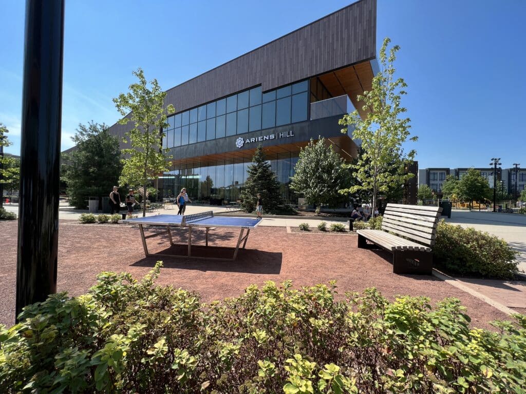 A brown triangular-shaped building is shown against a blue sky. In the foreground, a red brick patio holds a ping-pong table and a bench. Greenery surrounds the free ping-pong table in Titletown. 