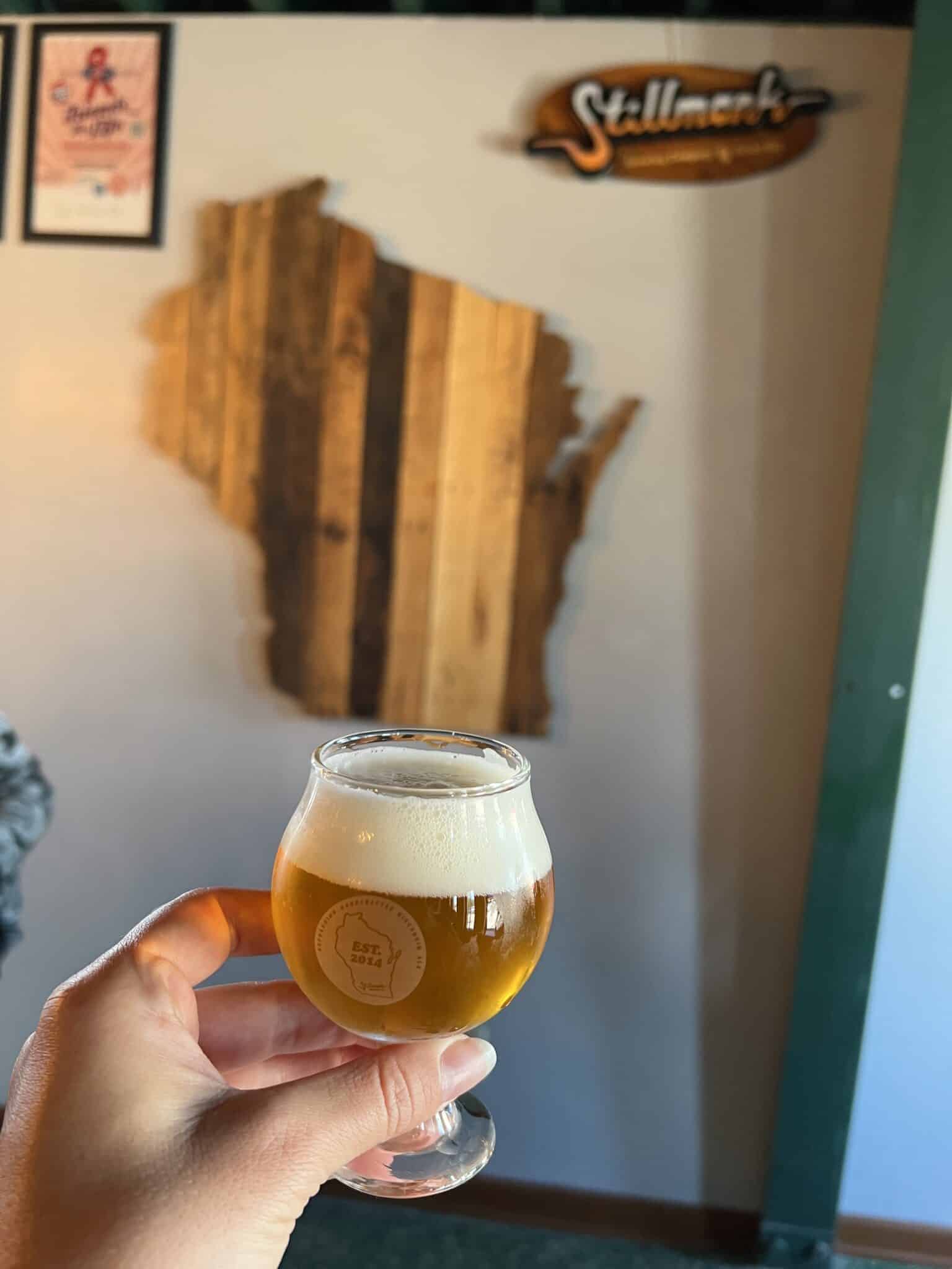 A hand holds a small beer goblet with a wooden Wisconsin-shaped sign in the background. Another sign in the upper corner says Stillmark.