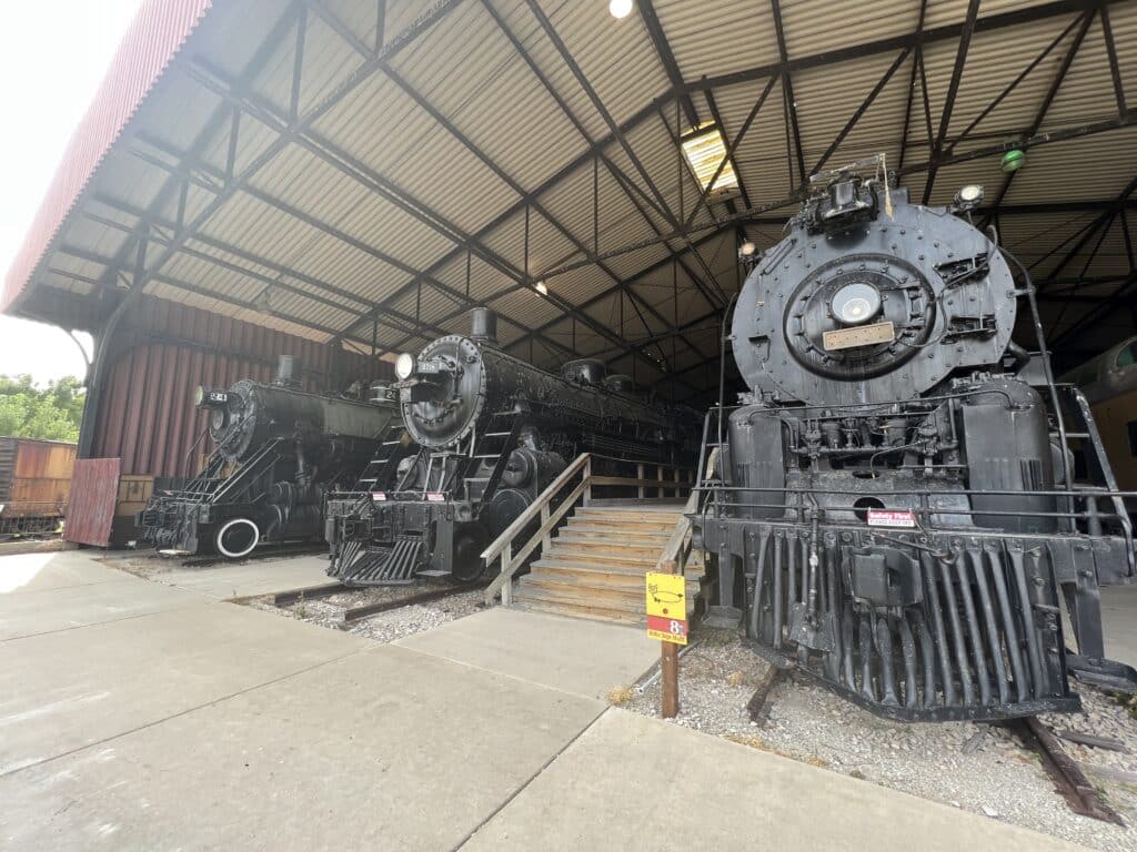 Three older black train steam engines are showcased under an awning with wooden stairs leading up to each. The trains are part of the National Railroad Museum in Ashwaubenon, near Green Bay. 