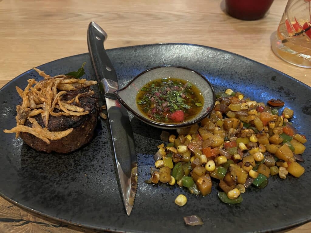 A pewter plate holds a buffalo steak with a side of grilled mixed vegetables. The steak is topped with fried onions and plated beside a ramekin of sauce and a large steak knife. 