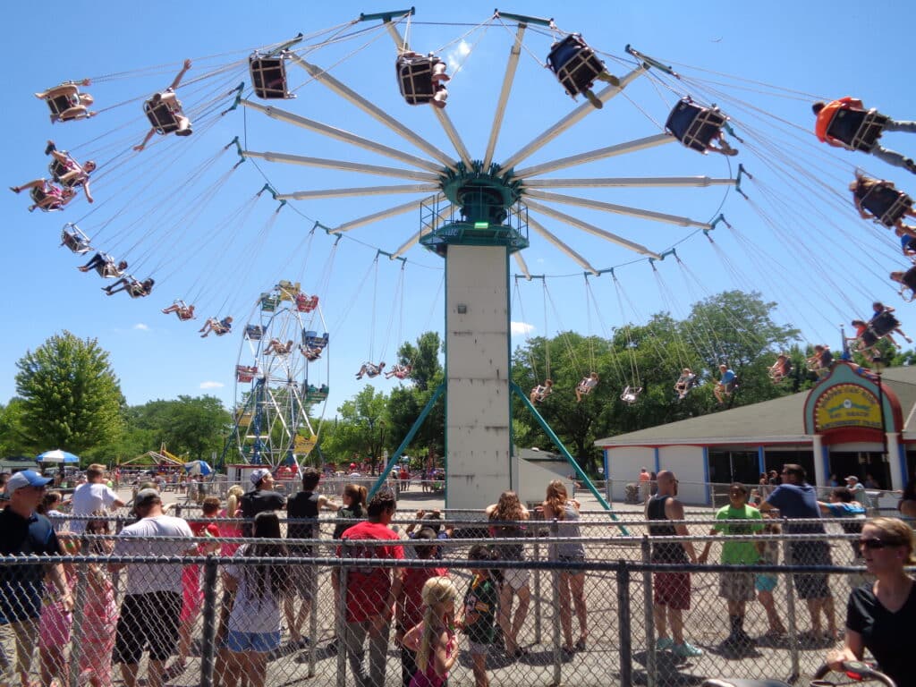 Families with children are waiting behind a chain-link fence in line to ride a suspended swing ride at the Bay Beach Amusement Park. Riders on the white swings spin in a circle, some with their arms outstretched against the blue sky above. In the background, there is a Ferris wheel and a refreshment building. 