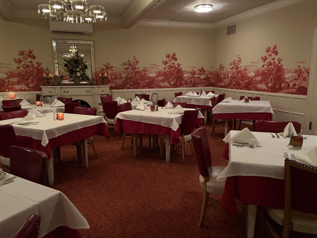 A classic supper club dining room at the Union Hotel in De Pere, WI, is decorated in shades of maroon with white table clothes. The walls feature a floral mural, and the square tables have a nod to the art deco style. 