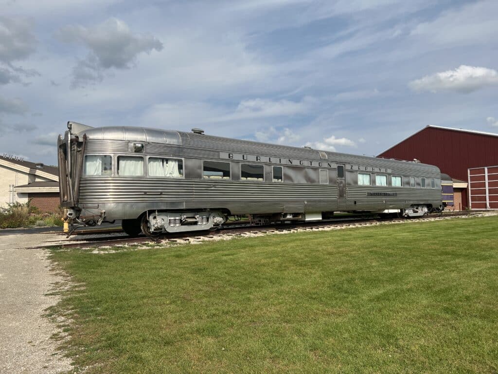 A silver mid-century passenger train car called the Burlington Zephyr is displayed on a track, surrounded by grass with a red bar-like structure in the background at Green Bay’s National Railroad Museum.  