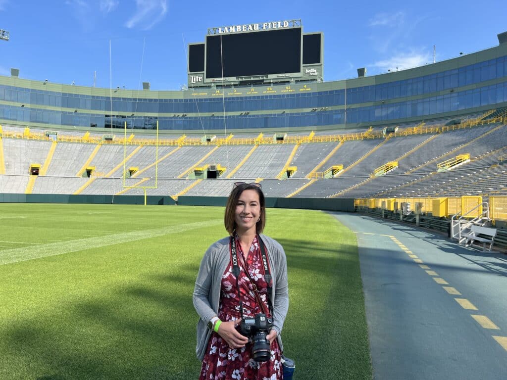Lindsey, a brunette woman in a burgundy floral dress and a grey sweater, is holding a camera and standing in the middle of Lambeau field. The stadium is empty behind her, the sky is blue, and the Lambeau field scoreboard is in the background.