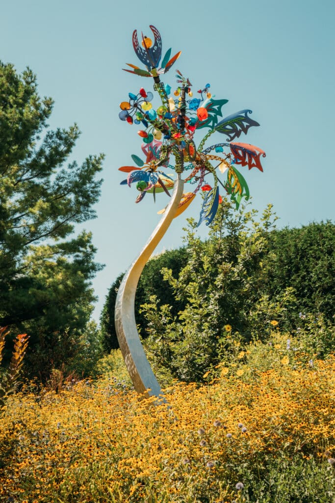 A colorful, whimsical sculpture is shown in a field of yellow Black-Eyed Susans and greenery. The sculpture resembles a tree with a silver trunk and colorful palms extending at the top in bright, primary colors. 