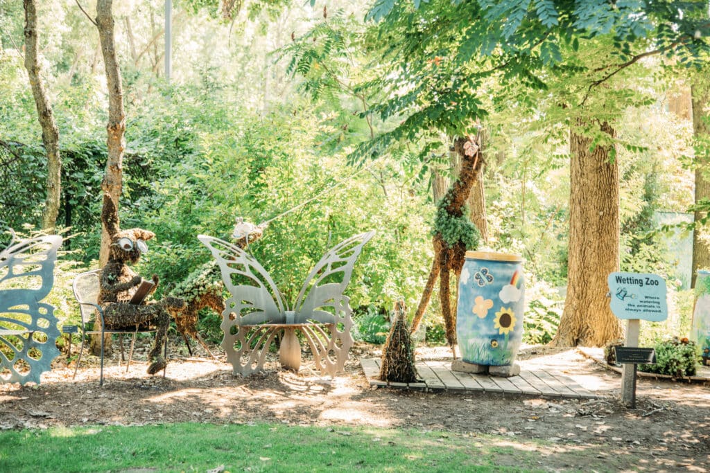 An outdoor garden display shows a rabbit made of moss wearing glasses and sitting in a chair. A giraffe, also made of moss, is positioned to eat from the trees. Butterfly-shaped silver benches and a colorfully painted rain barrel also sit within the "Wetting Zoo." 