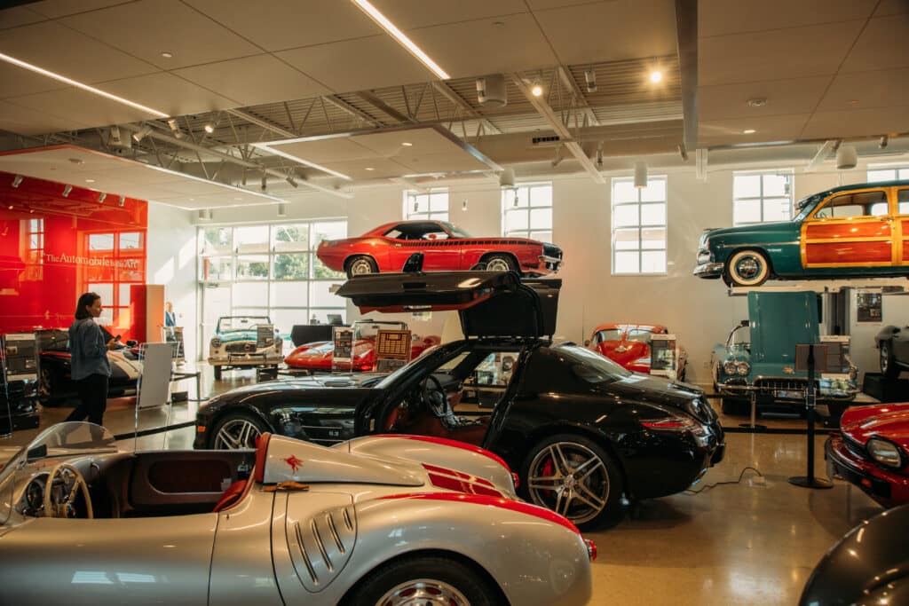 A showroom of vintage luxury cars is brightly lit. The colorful cars include silver, black, and a red car on an elevated platform. 