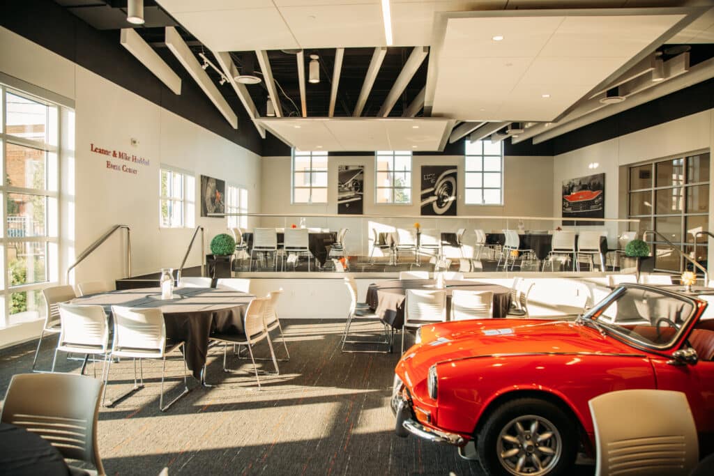 A dining and meeting space is surrounded by windows and bright white walls in the Automobile Gallery & Event Center in Green Bay, WI. There is a red classic car in the foreground with café tables and chairs filling the room.