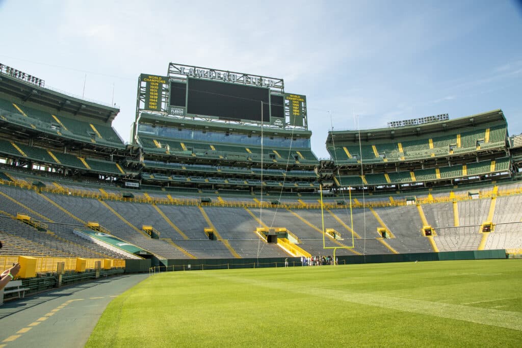 An image of empty Lambeau Field Stadium from the field. The grass is green, and the sky is blue. The grey stadium seats are shown beneath the marquee. 
