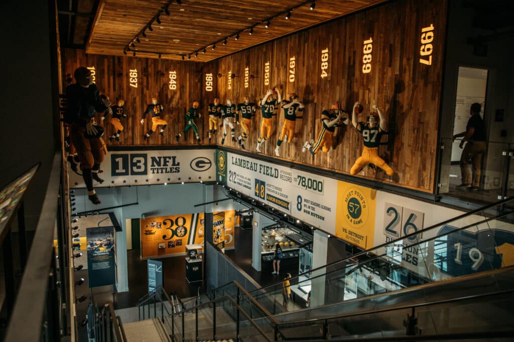 A staircase landing at the Green Bay Packers Hall of Fame features football player mannequins wearing yellow pants and green jerseys, suspended in alcoves. Green Bay Packers memorabilia adorns the walls with white signage and green and yellow lettering. 