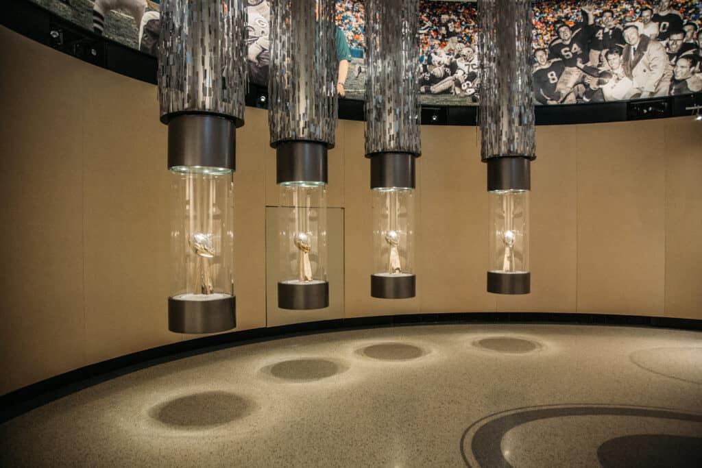 Four suspended cylindrical glass trophy cases contain Super Bowl trophies. The Vince Lombardi trophies feature a silver football on a pyramid-shaped stand. 