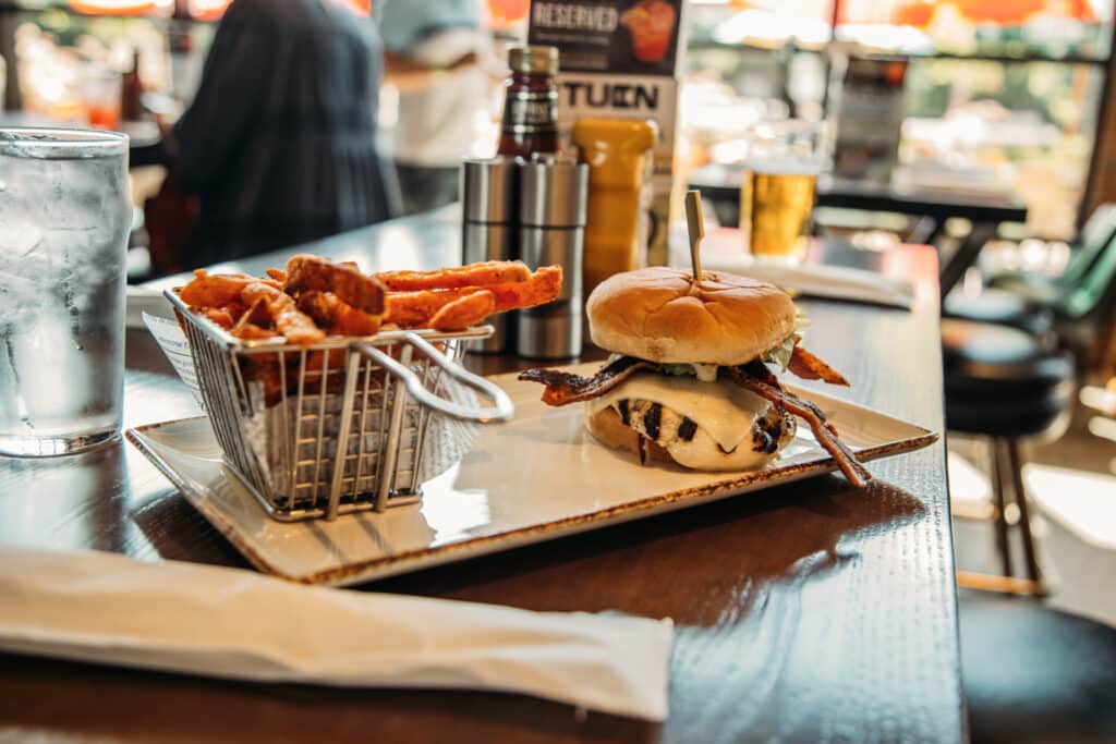 A silver basket of French fries is plated alongside a bacon-grilled chicken burger topped with Swiss cheese. The square plate rests on a bar counter, with a glass of ice water and other beverages in the background.  