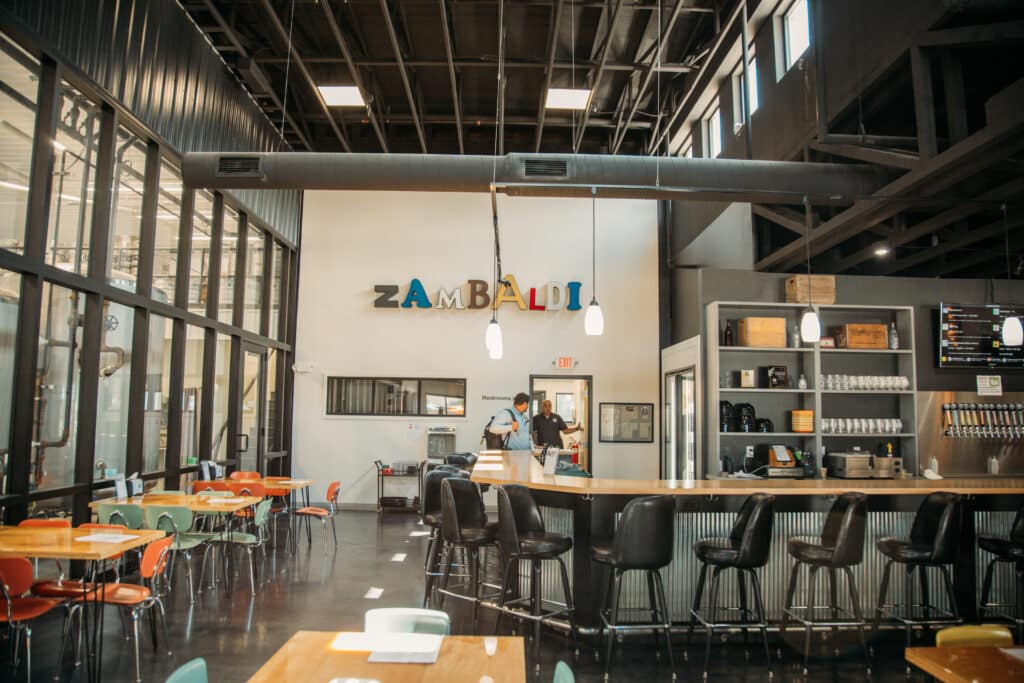 An industrial-style brewery bar has a wooden countertop and classroom-style chairs in colorful oranges, greens, and pewter. A sign against a white wall says Zambaldi in various colors and typefaces. 