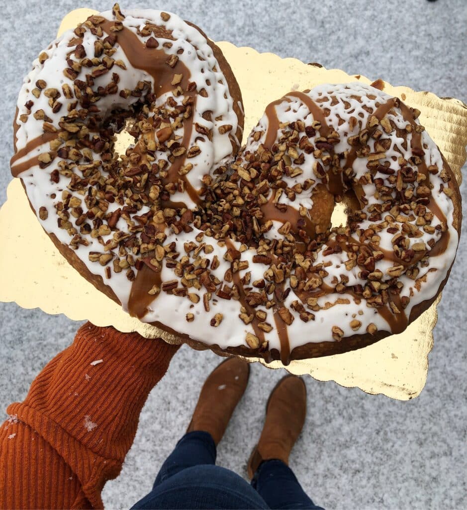 A large, pretzel-shaped kringle pastry is covered with white frosting, caramel, and nuts. A person in a rust-colored sweater and brown boots is holding the kringle atop a gold foil tray. 