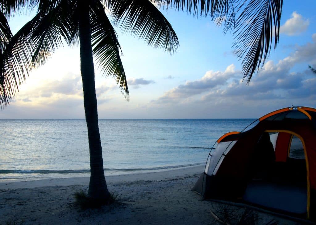 A large camping tent pitched right on the shoreline of a tropical Caribbean beach at dusk, overlooking the calm waters and a clear sky.