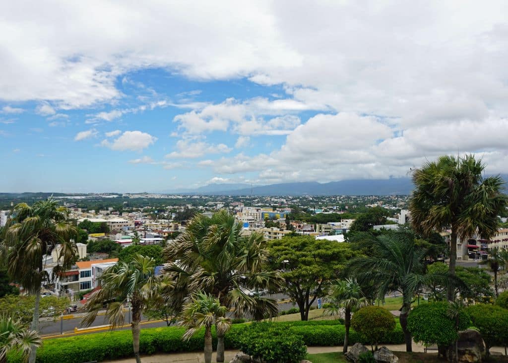 A landscape image of the Dominican Republic -- tropical trees in the foreground, and an expansive residential city spreading out in the distance, under blue skies and white clouds.