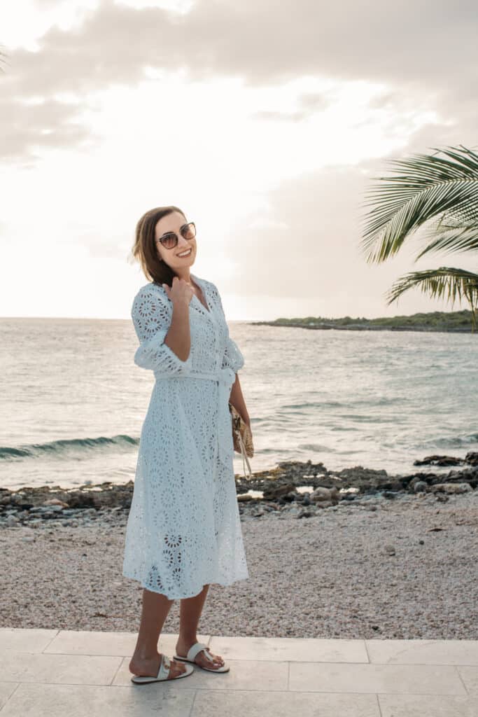 Lindsey of Have Clothes, Will Travel wearing a white eyelet midi dress from Lilly Pulitzer, standing by a stony beach at sunset in Curacao