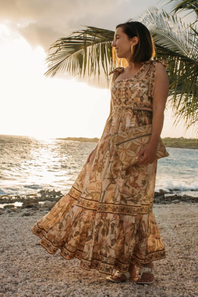 The rocky beach at Sandals Curacao, and Lindsey of Have clothes, will travel wearing a Zimmermann maxi dress and sandals standing by a palm tree at sunset.