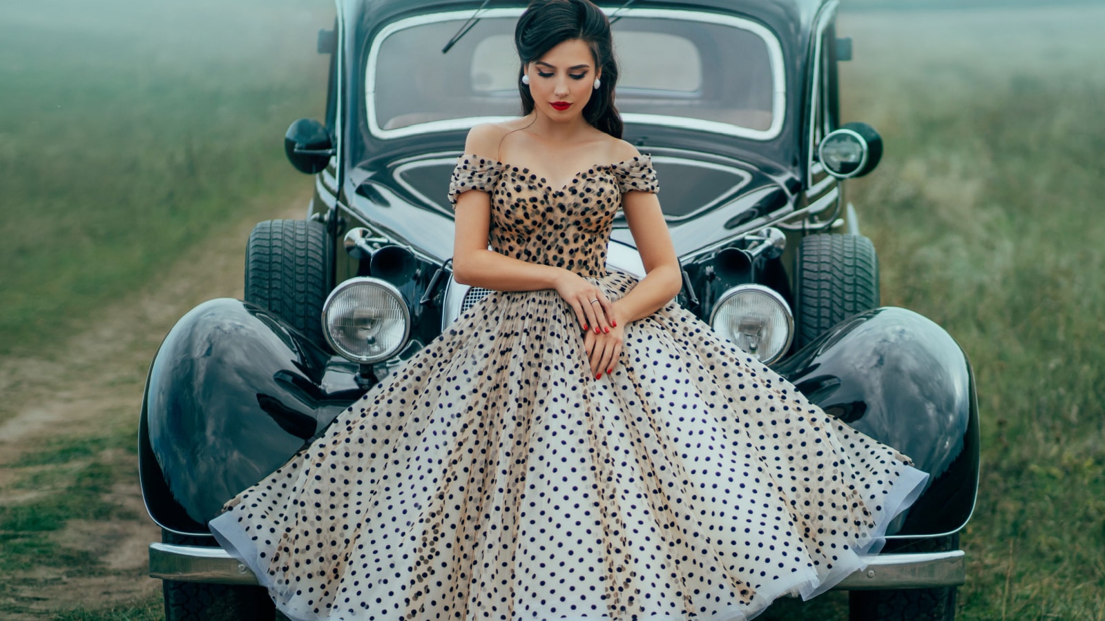 Young beautiful sexy woman in pin up style clothes posing near black retro car. Polka dot white dress, vintage hairstyle, red high heels. Background road green nature fog. Girl fashion model driver