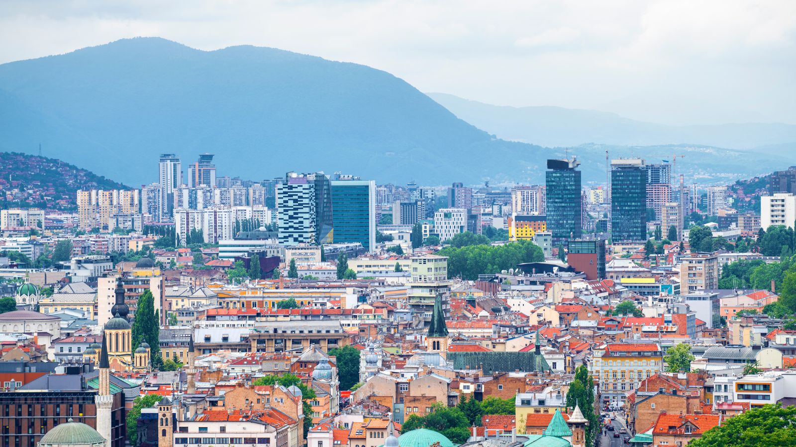 An aerial landscape view of the city of Sarajevo, BiH. Smaller colorful homes and building in the foreground are followed by taller, modern buildings, with vast mountains in the background.