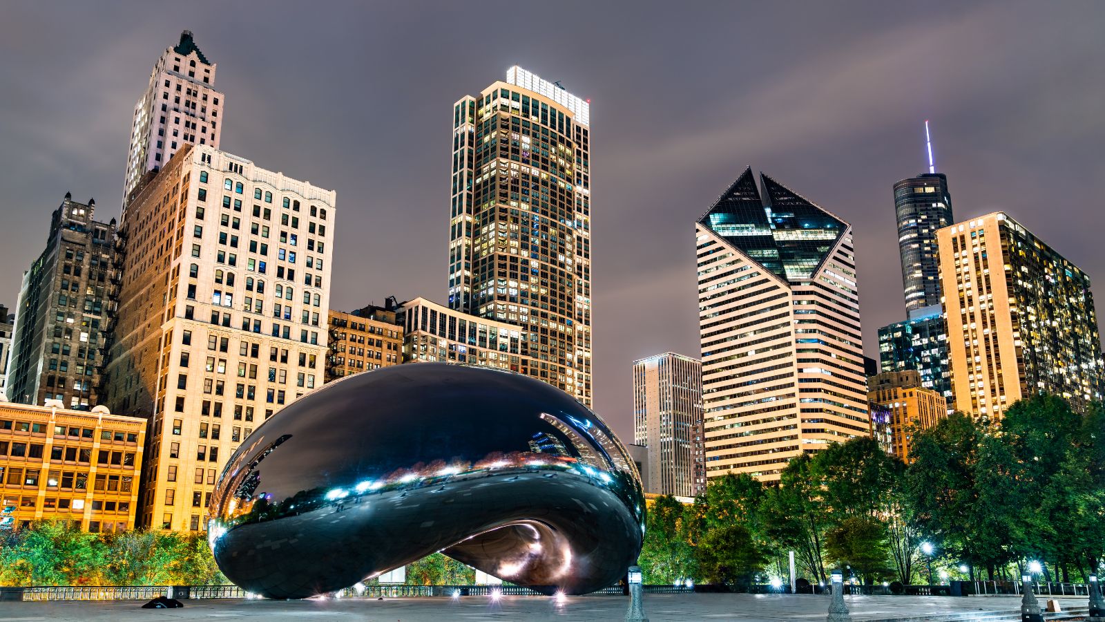 The Bean sculpture in Millennium Park in the middle of downtown Chicago, surrounded by skyscrapers lit up against a cloudy night sky.