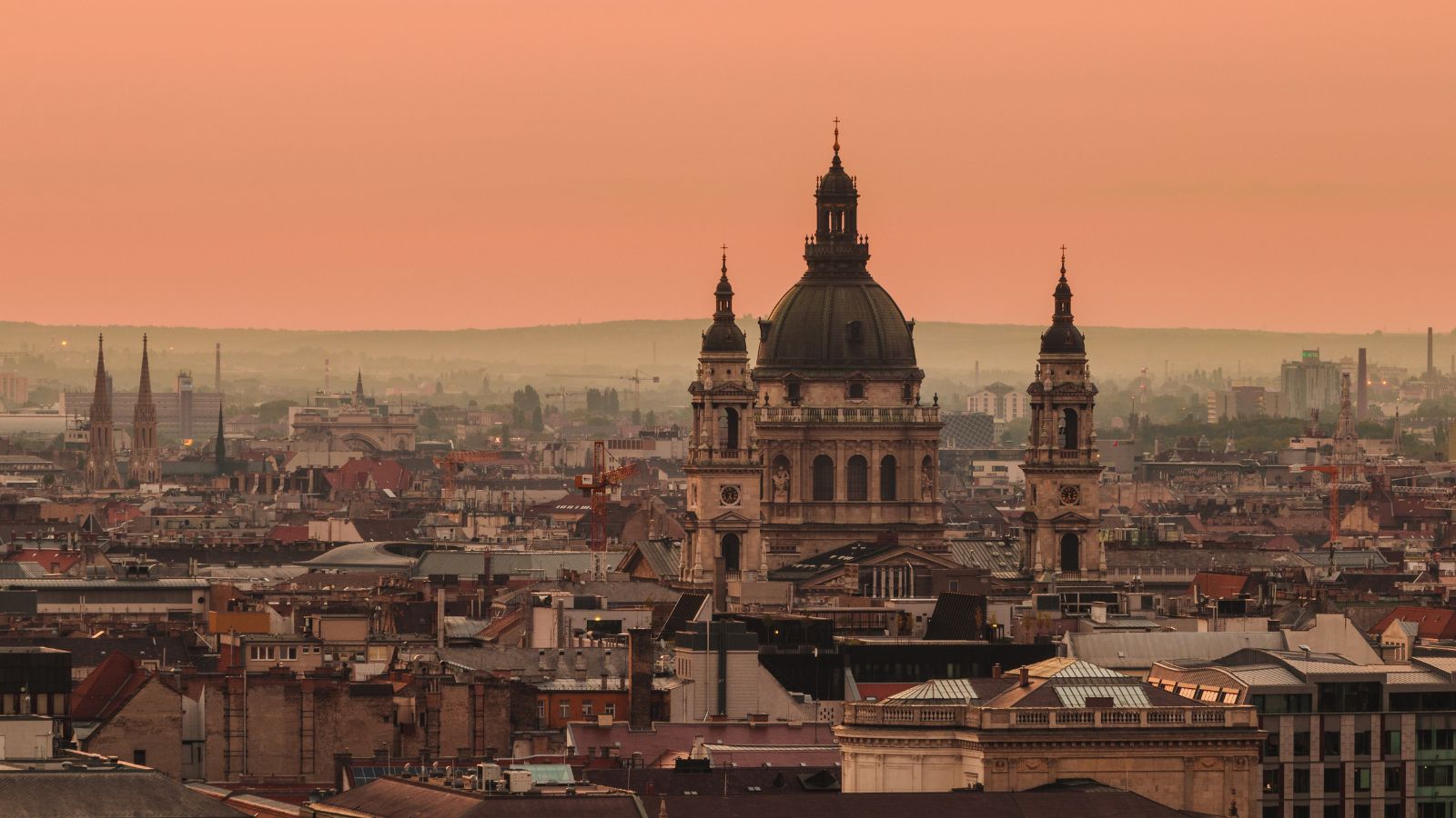 An aerial shot of the city of Budapest, Hungary at dusk. An orange sky looms over ancient buildings, with St. Stephen's Basilica in the center.