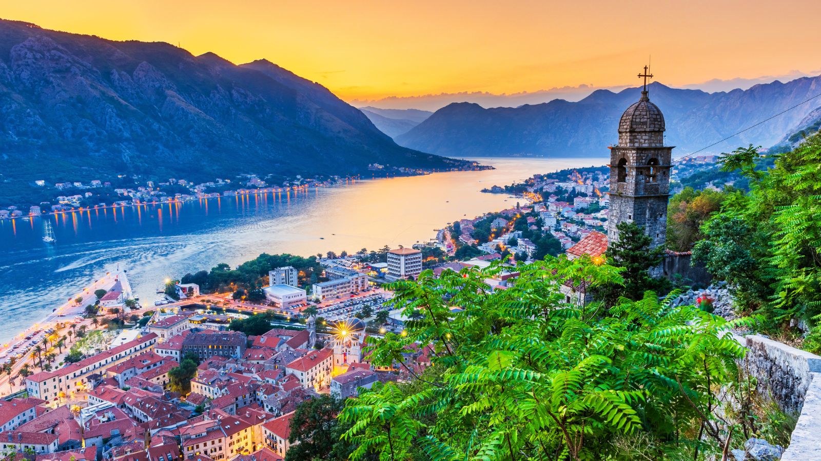 A sunning aerial view of Kotor Crna Gora at sunset. The winding waters weave through mountain ranges on one side, and a bustling city center on the other side, packed with buildings and green trees.