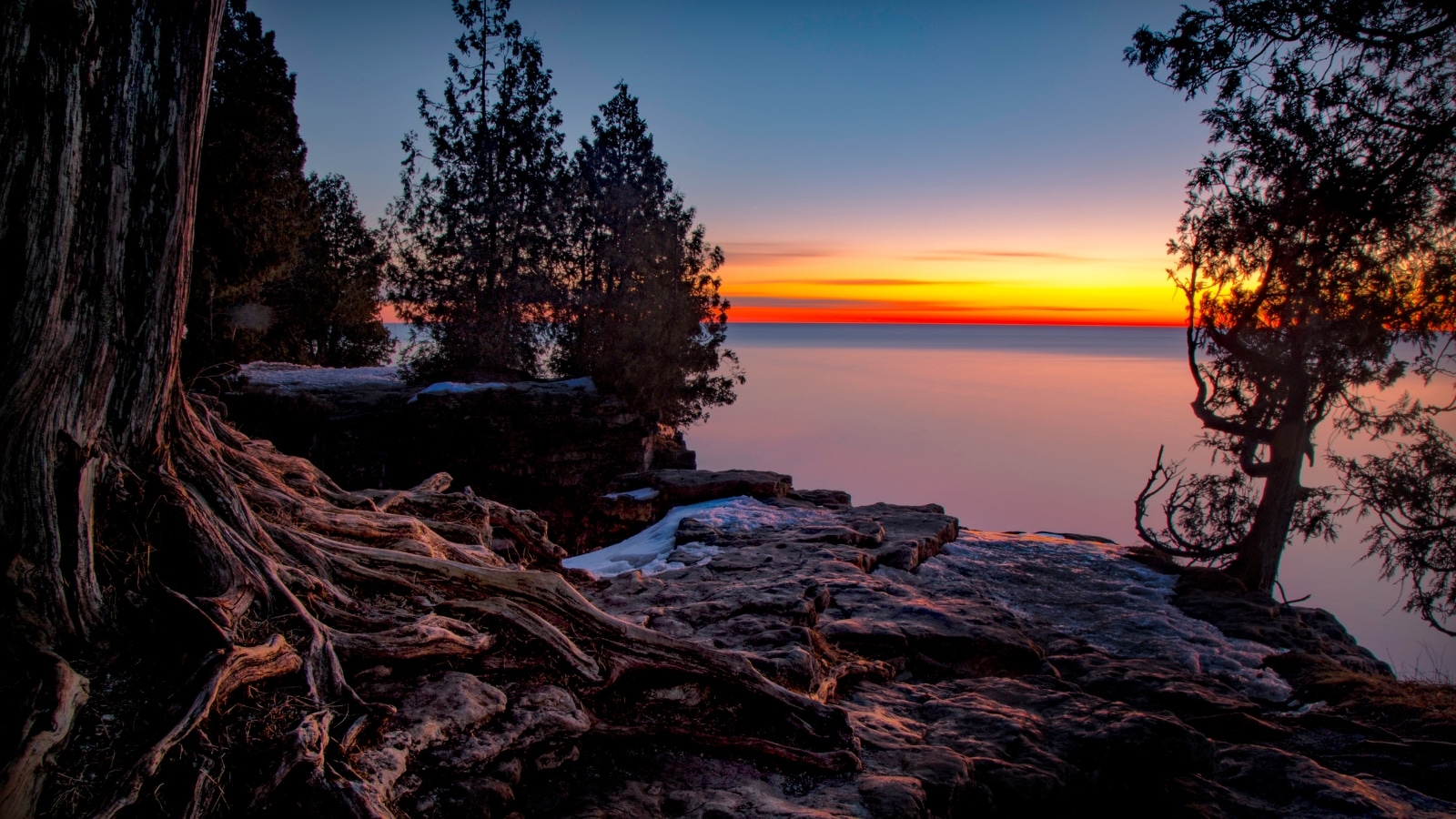 A Door County, Wisconsin shoreline at dusk. The rocky, tree-lined shoreline looks out over the lake to the sun setting in a colorful sky.