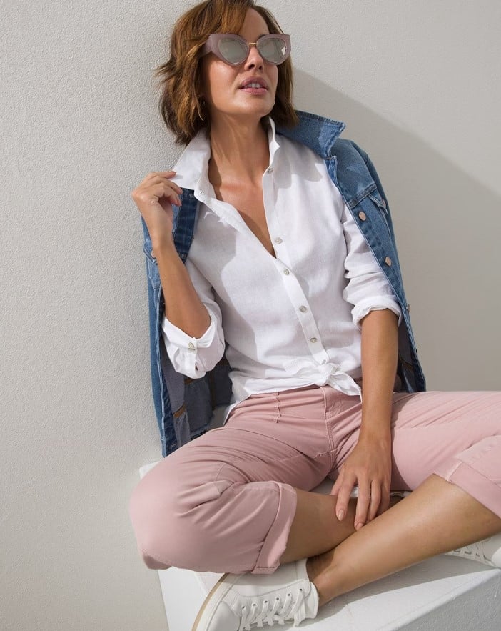 Woman sitting cross legged, wearing sunglasses a white button down paired with pink cropped pants and a jean jacket