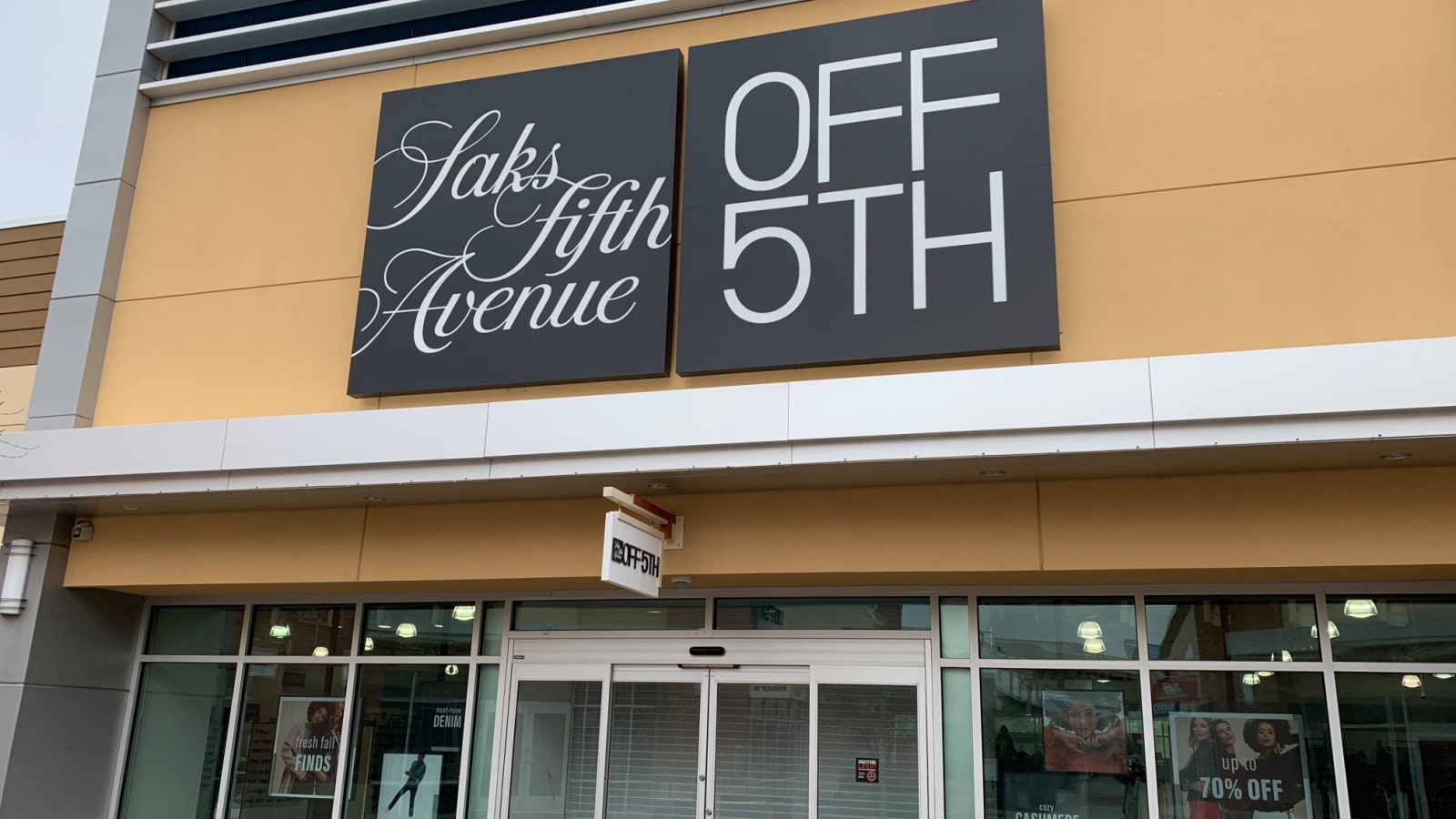 The storefront of a Saks Off Fifth store features two large signs hanging over the doorway. One says "Saks Fifth Avenue" in decorative cursive font; the other says "Off 5th" in large, modern typeface.