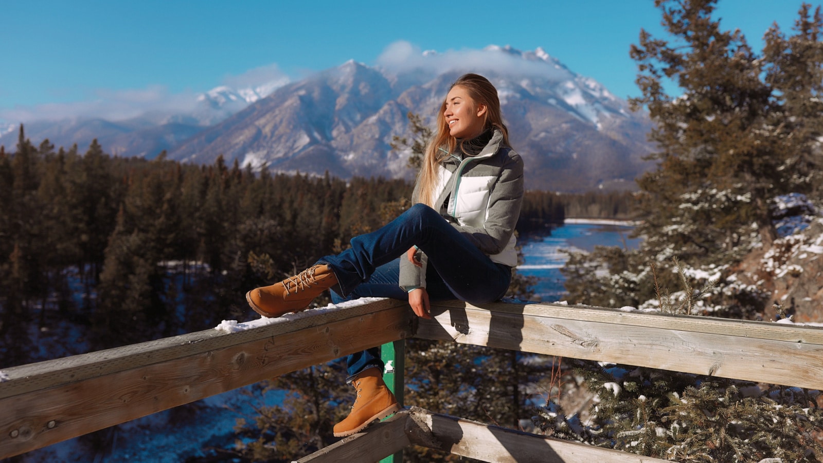 Pretty lady wearing winter fashion and comfortable clothes to travel around cold and snowy Canada. sitting on a wooden fence, looking far away, catching the sun light on her tender face. Picturesque