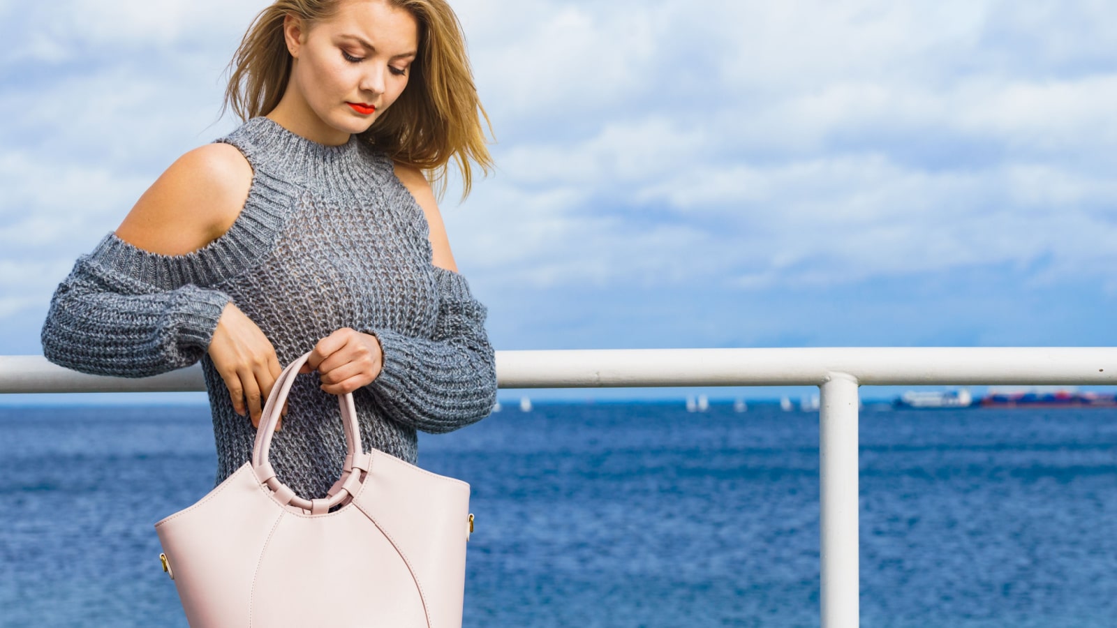 Attractive fashion woman in trendy outfit long sleeve cold open shoulder pullover outdoor on sea coast. Female model holding handbag.