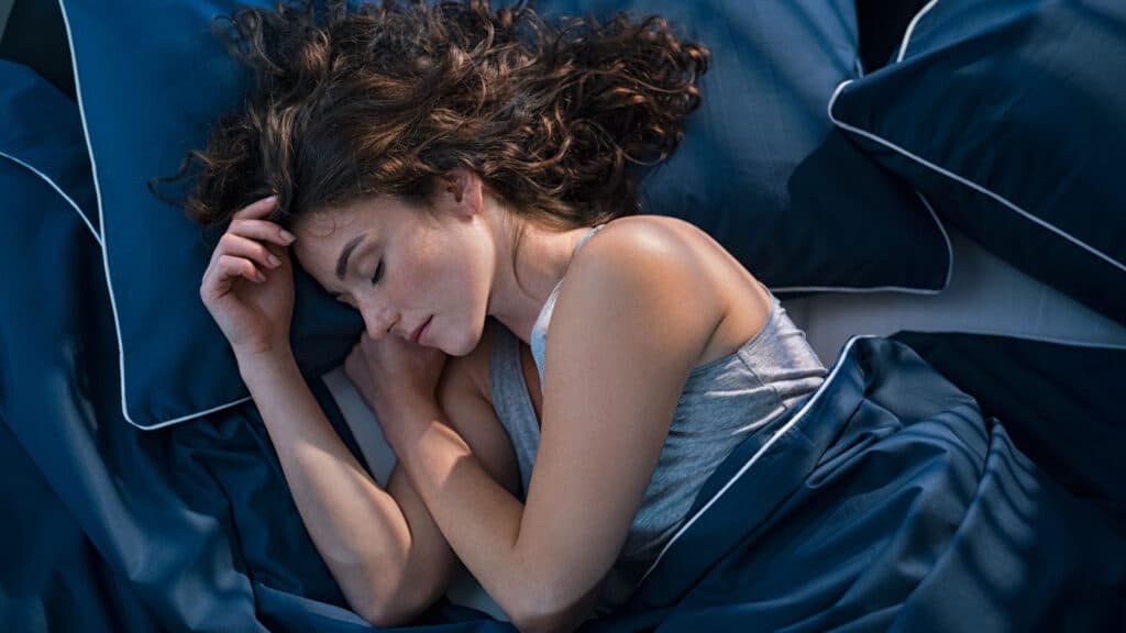 Top view of young woman sleeping on side in her bed at night. Beautiful girl sleeping profoundly and dreaming at home with blue blanket. High angle view of woman asleep with closed eyes.