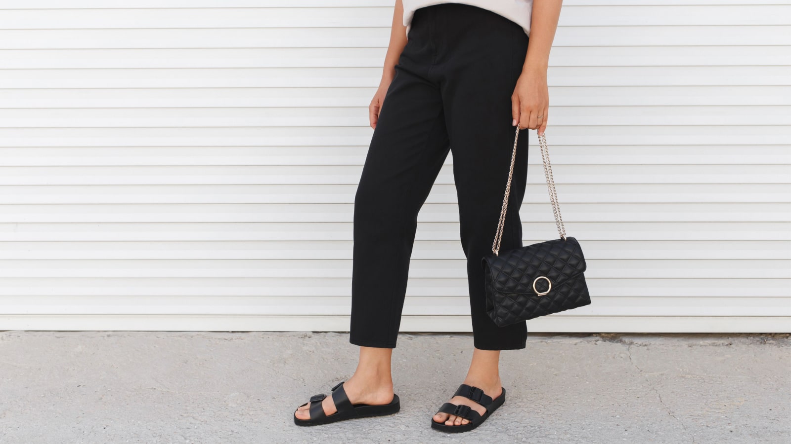 Woman wearing beige t-shirt, black pants, bag and flat sandals walking outdoor near white roller door. Details of stylish trendy basic minimalistic casual outfit. Street fashion. Women's legs, no face Birkenstock