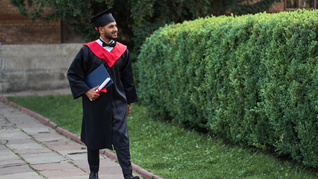 Handsome indian graduate walking in university campus in graduation glow and diploma, copy space