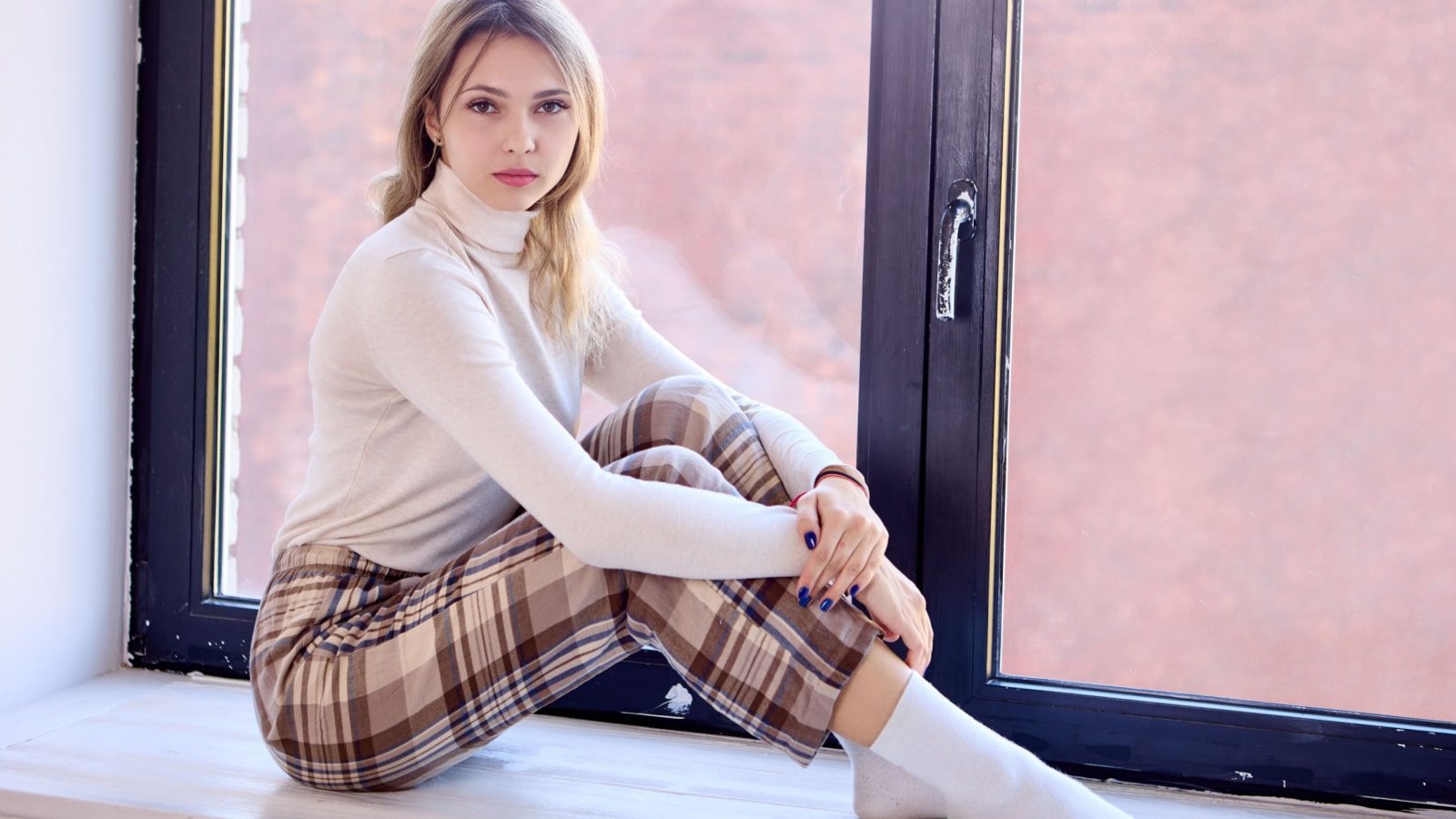Woman in plaid pants is 24 years old, sitting on windowsill.