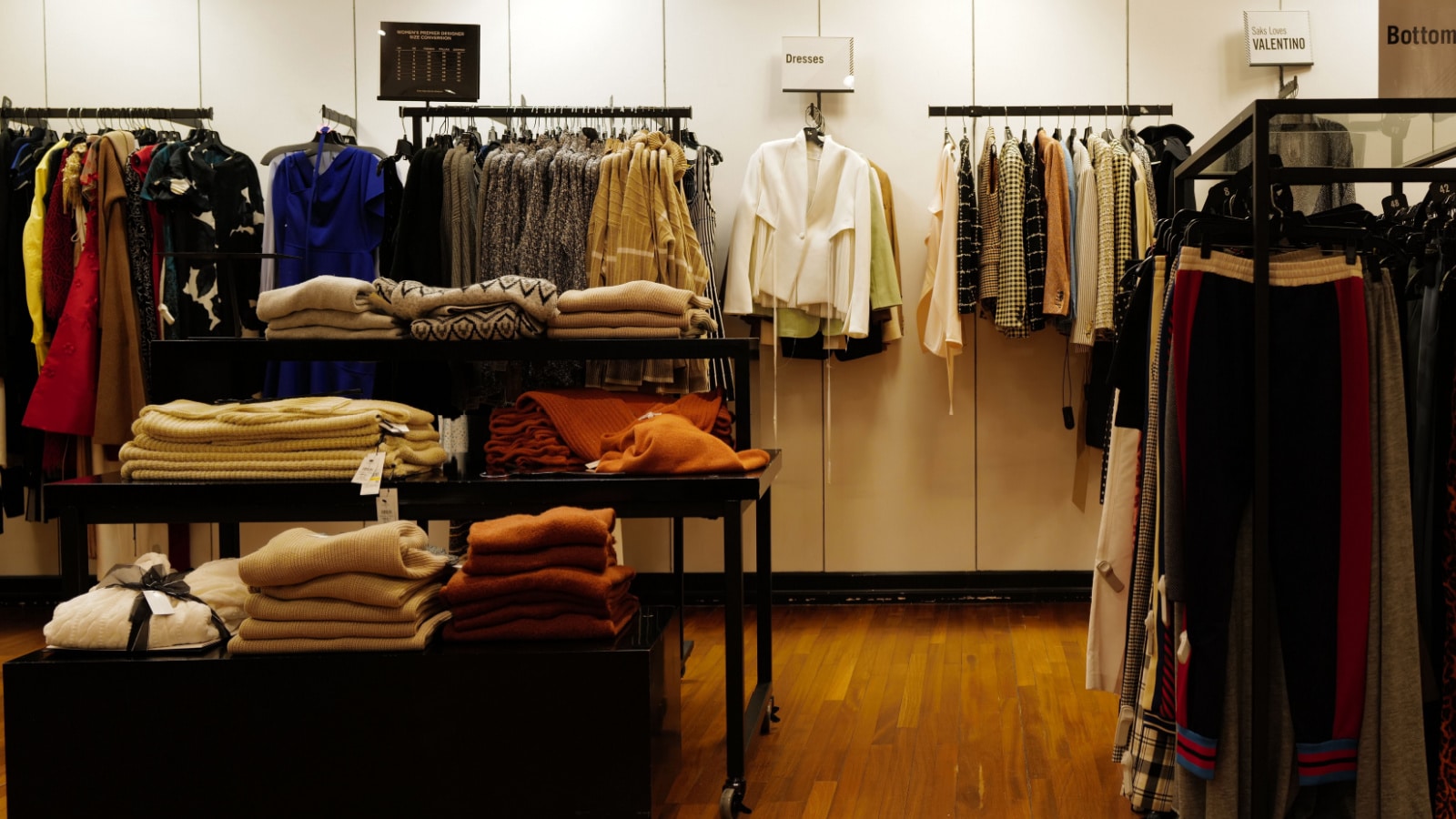 A retail store clothing display at Saks Off Fifth features racks of sweaters, tops, pants, and accessories in a warmly-lit department store.