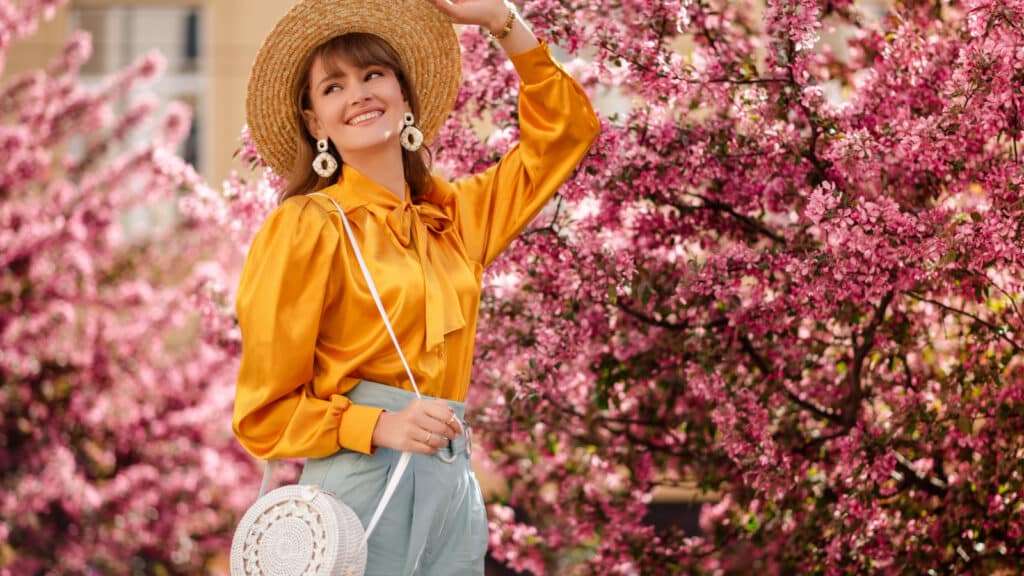 Outdoor lifestyle portrait of elegant happy smiling woman wearing trendy straw hat, yellow satin blouse, blue trousers, with shoulder wicker round bag, walking in street near pink spring blossom trees
