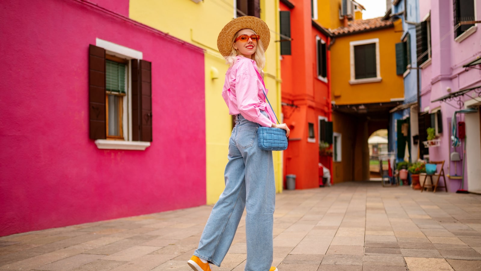 Happy smiling female traveler wearing stylish hat, glasses, pink shirt, wide leg trousers, walking, posing near colorful houses in street. Travel, tourism, vacation, fashion, lifestyle conception