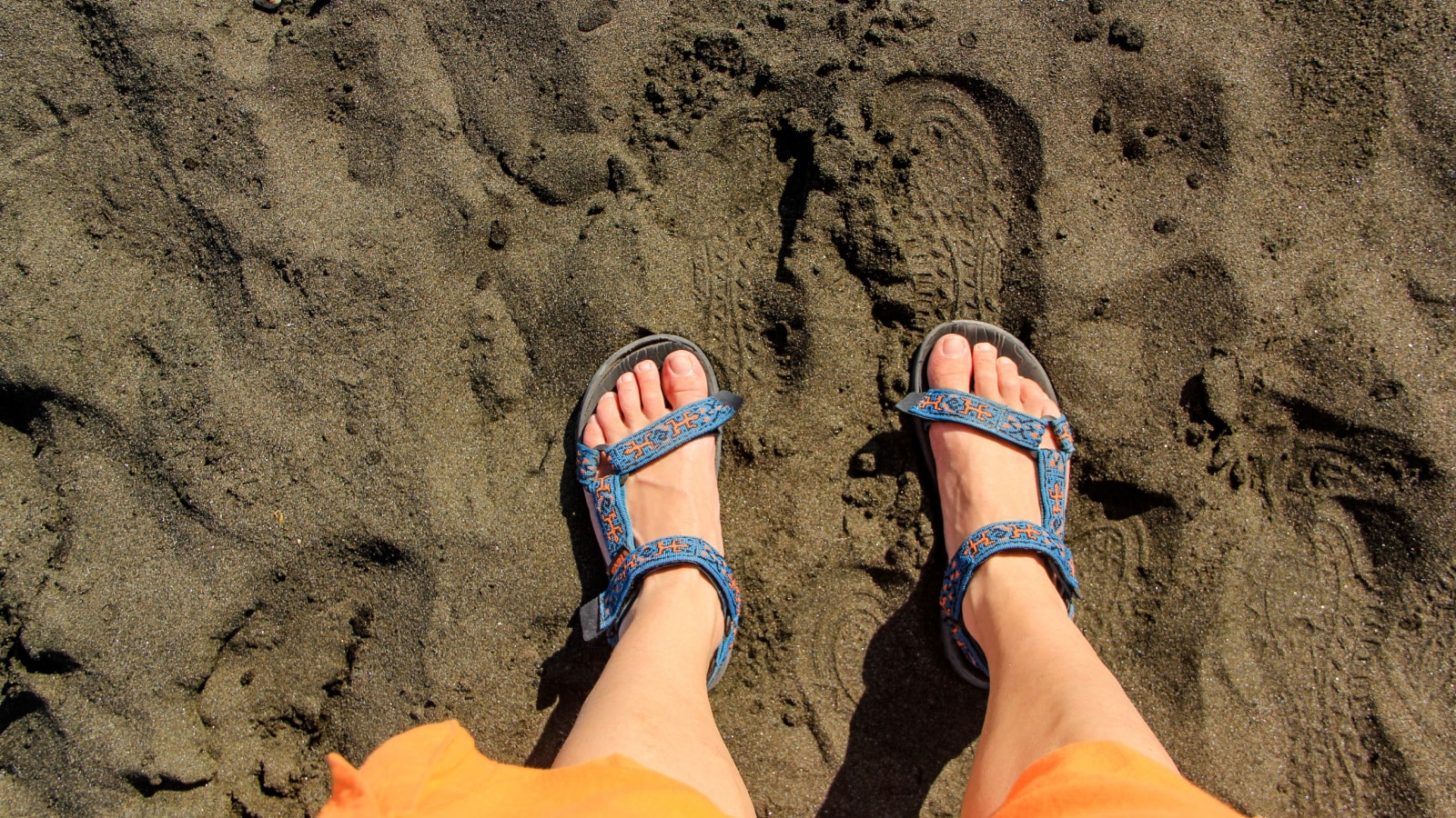 Legs in blue Teva sandals and orange breeches against the backdrop of black sand on the seashore in Naples